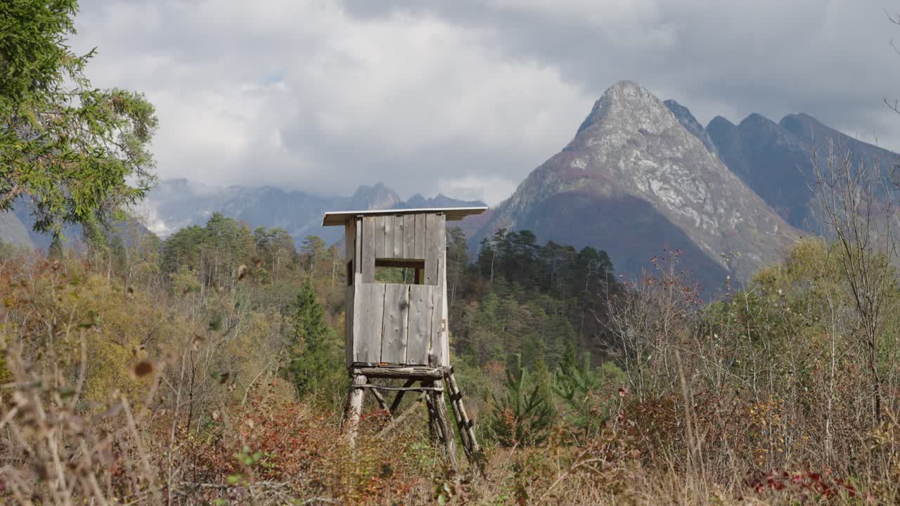 Høyt jaktvakttårn i tre står over våtmarker med fjellskog i bakgrunnen på en overskyet dag