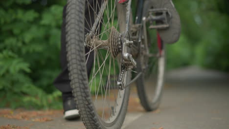 close-up of someone rotating the pedal of their bicycle backwards with their foot on a paved path, the setting features blurred greenery in the background