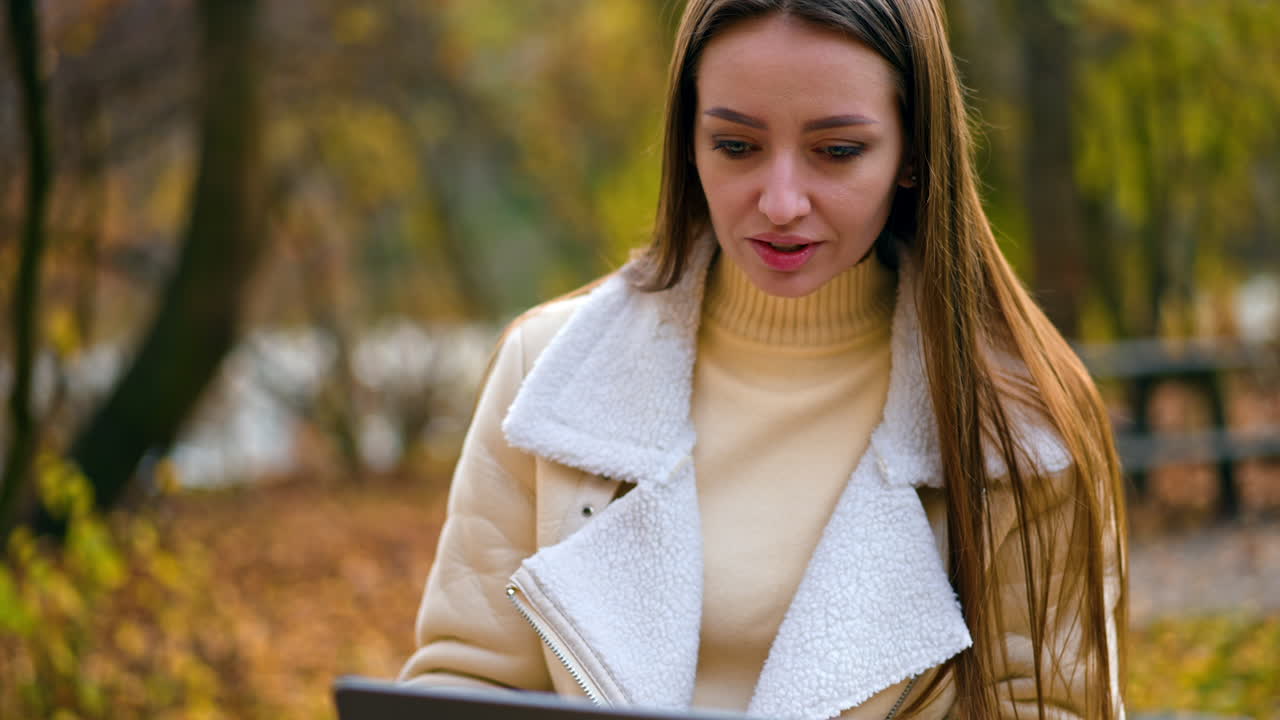 Modern young woman using laptop sitting in the nature. Brunette beautiful lady sitting on the table looking at computer screen. Autumn nature backdrop.