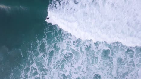 an aerial from above of waves frothing below, before a surfer comes into focus as they catch a wave on the california coast