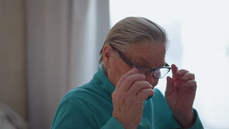 elderly woman in teal green sweater sitting on bed, adjusting her glasses with focused expression, soft natural light filtering through the window, calm and peaceful indoor environment with minimalistic decor