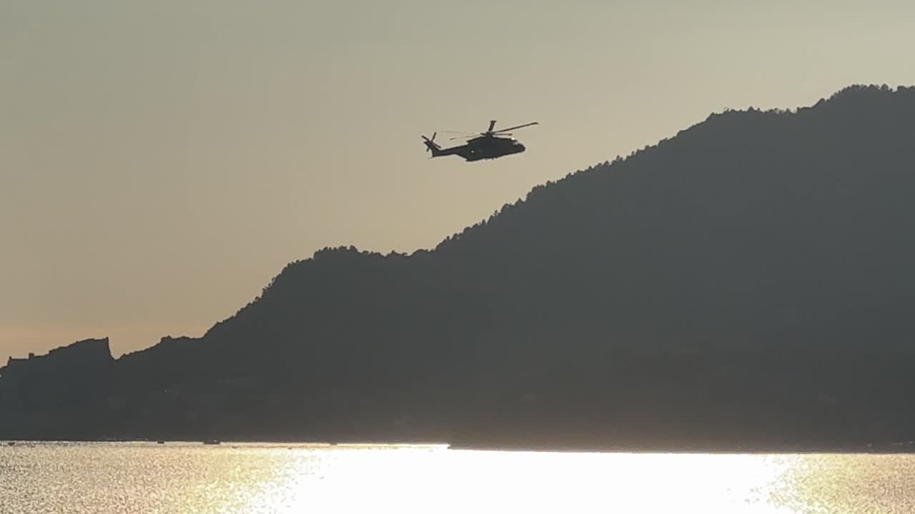 Silhouette of an Italian Navy helicopter flying over the sparkling sea at sunset near coastal mountains in the Gulf of La Spezia, Italy
