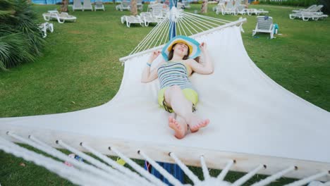 una chica brillante con ropa de colores frescos y un sombrero de borde ancho está descansando en la costa en una hamaca de textil blanco