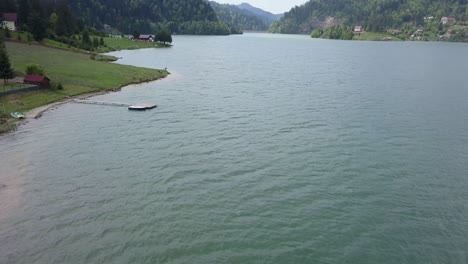 Aerial-Panning-Shot-of-edge-of-lake-with-a-cabin-and-trees-in-the-background-and-a-pier-in-the-foreground