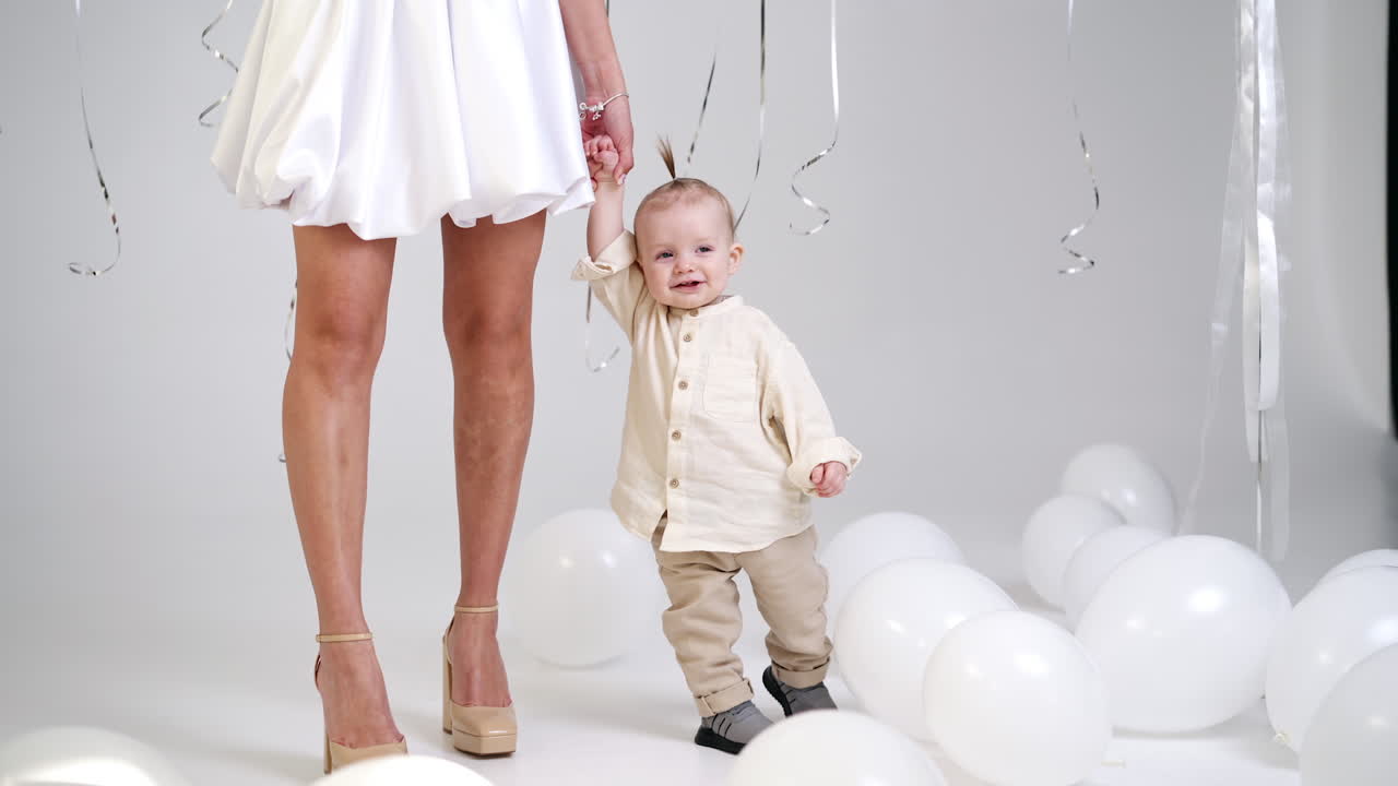 Unrecognized woman in short white dress holding her baby by the hand. Cute smiling child walks around his momma. Studio footage with white balloons around.