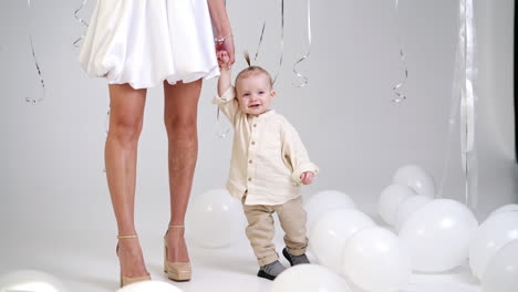 unrecognized woman in short white dress holding her baby by the hand. cute smiling child walks around his momma. studio footage with white balloons around.