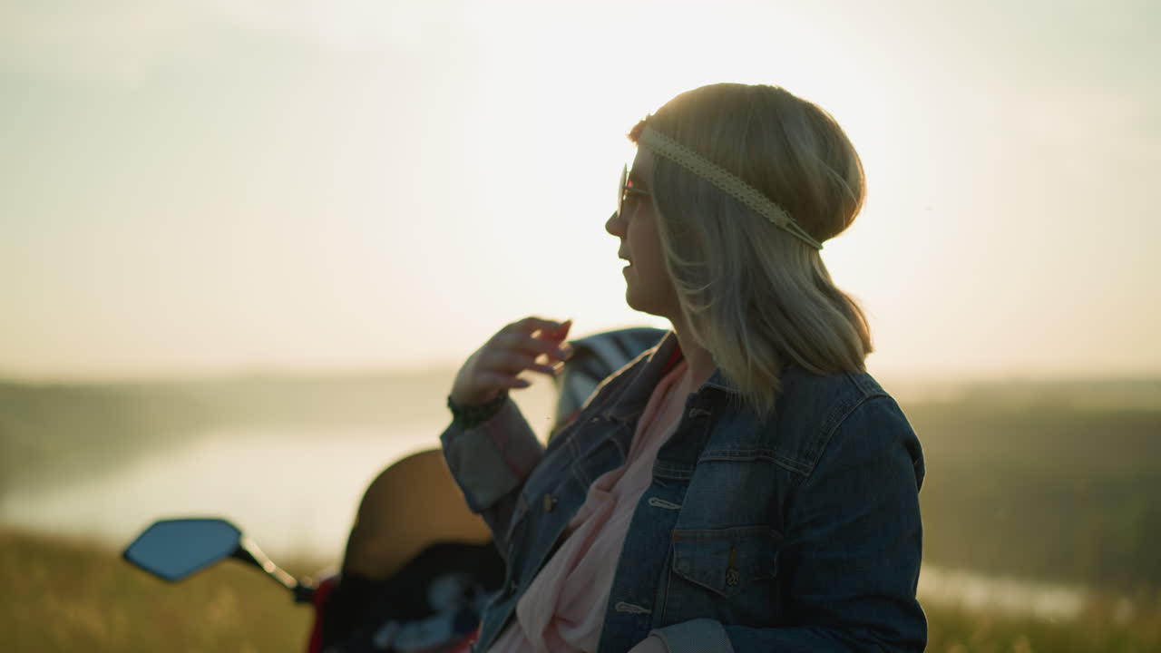 A woman adjusts her hair while wearing a headband and denim jacket, with the sun glowing behind her head