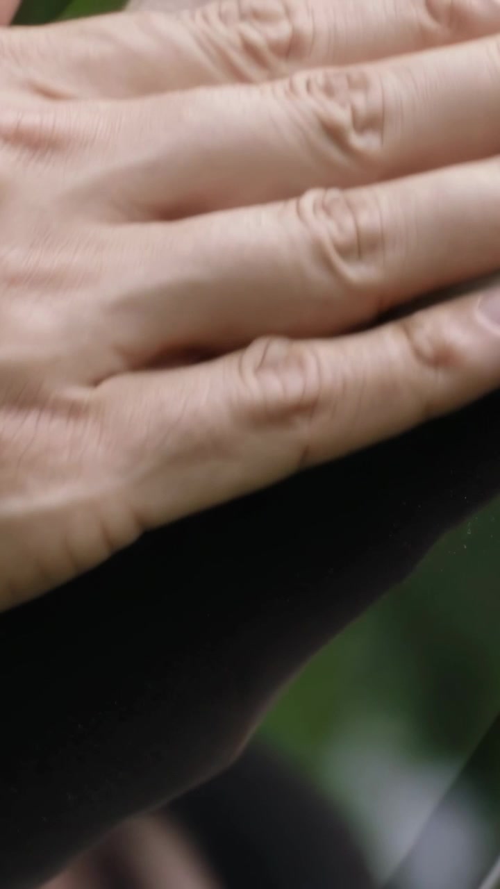 Vertical video: Noticing smudge, Asian man in collared shirt wiping tablet with napkin in foliage