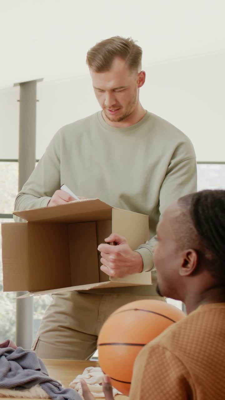 Vertical video: Picking up marker, man labeling box at home table for packing, with basketball