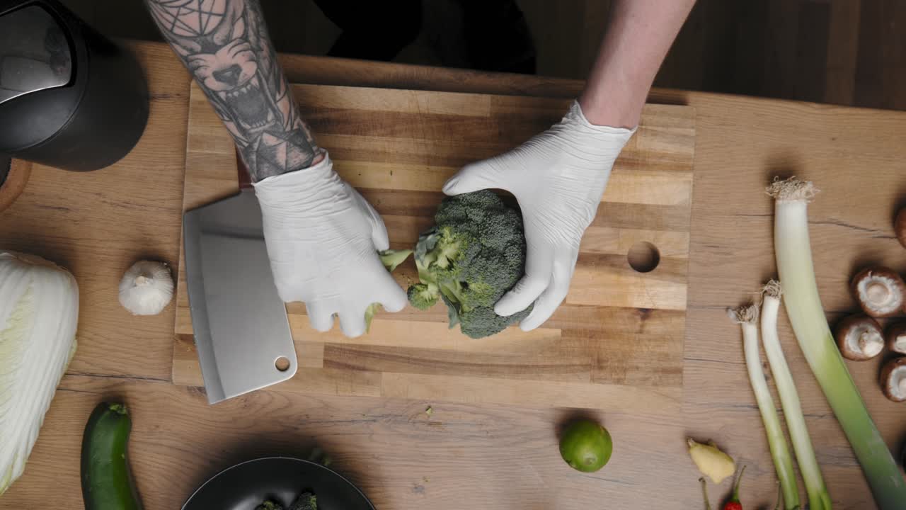 Fresh green broccoli being cut on a wooden board by young professional male chef in an elegant black shirt with tattoos