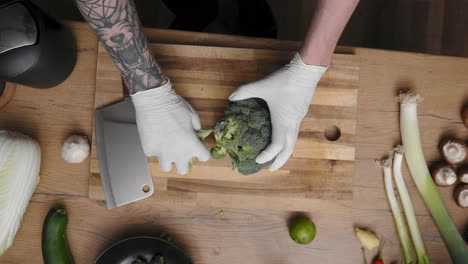 fresh green broccoli being cut on a wooden board by young professional male chef in an elegant black shirt with tattoos