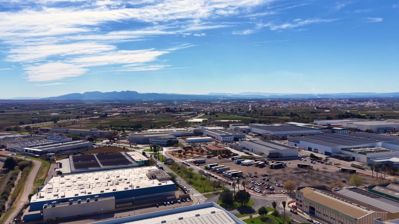 Vista dall'alto di un parco industriale spagnolo con magazzini, strutture logistiche e pannelli solari sui tetti, circondato da terreni agricoli e montagne sotto un cielo azzurro