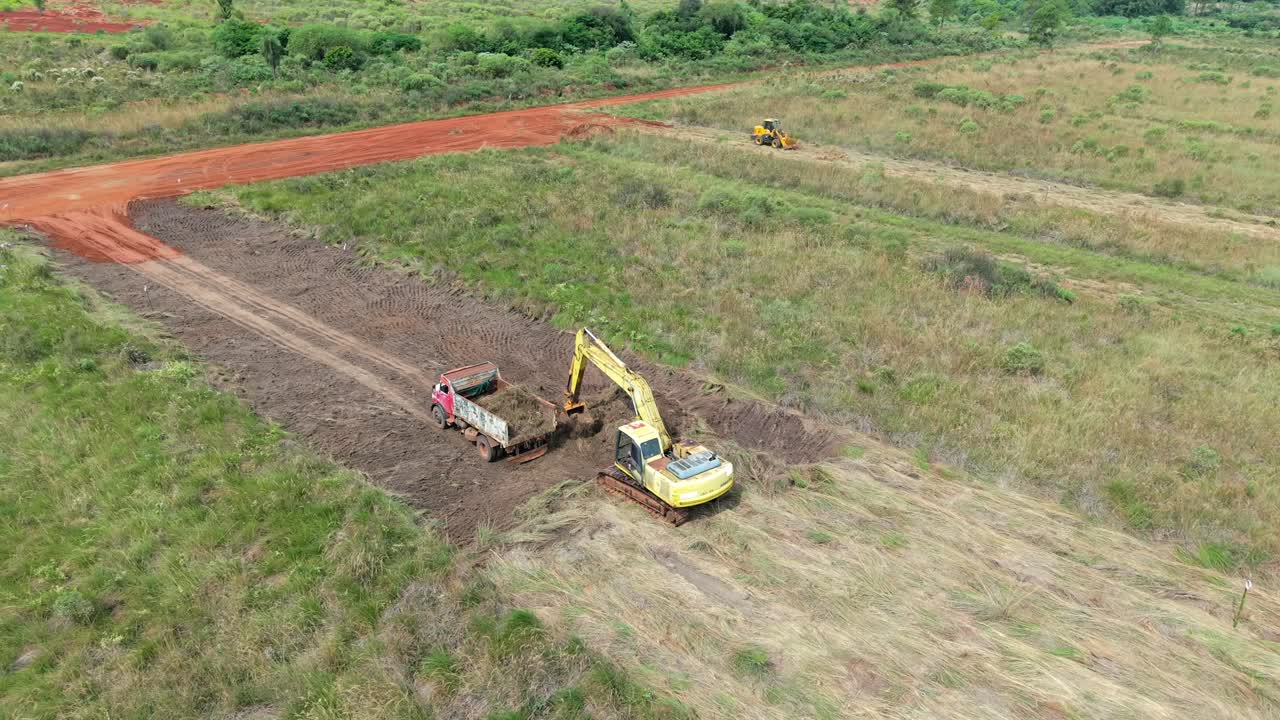 Yellow excavator loading truck on a rectangular farmland plot with red soil.