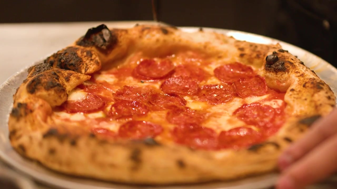 Primo piano di mani che usano una rotella tagliapizza in metallo per tagliare una pizza calda al salame piccante in cucina.