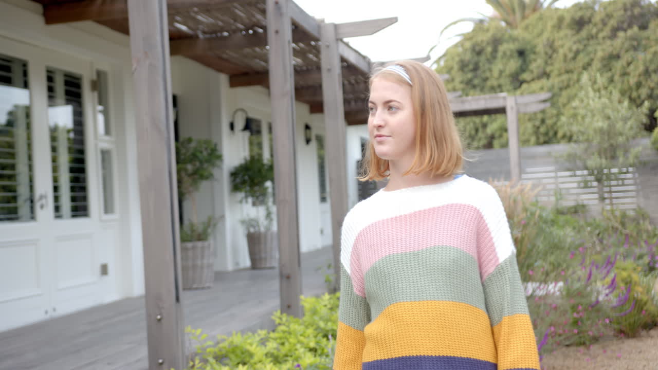 Woman in colorful sweater standing outside home, looking around garden area, copy space