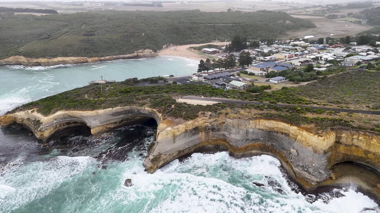 Drone footage captures the dramatic cliffs and ocean waves of Port Campbell, Australia, under overcast skies with a sweeping aerial perspective