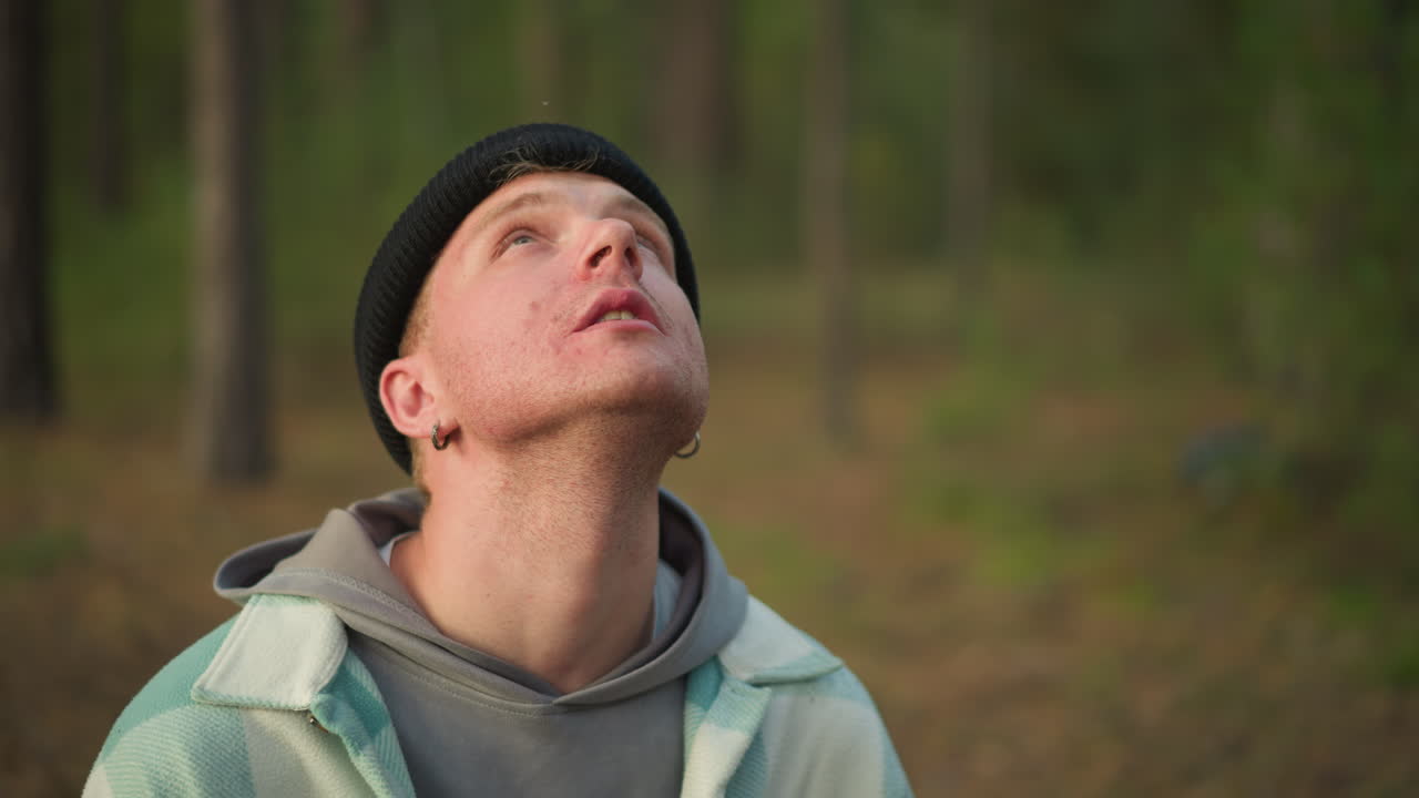 Giovane uomo nella foresta che sorseggia da una tazza di metallo, indossa un berretto di lana e una giacca a quadri, luce mattutina tra gli alberi di pino, escursionista in pausa sul sentiero, campeggiatore che respira vapore, viaggiatore che guarda il cielo