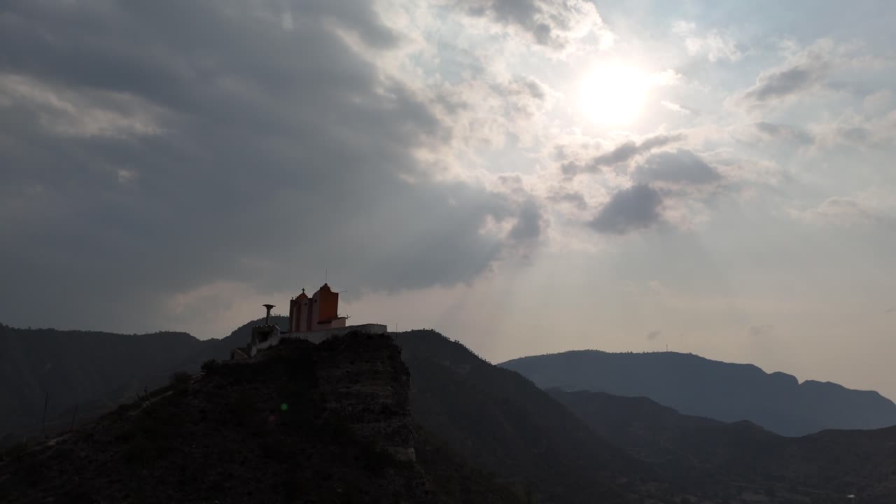 A drone flies around a church on a hill, capturing the sun shining through clouds in the sky.