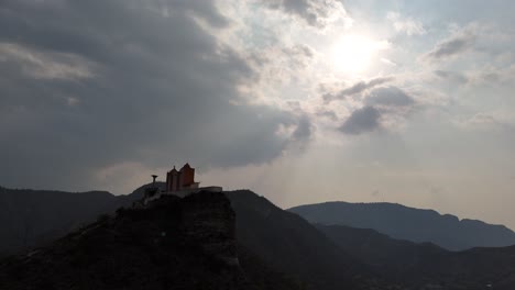 a drone flies around a church on a hill, capturing the sun shining through clouds in the sky.