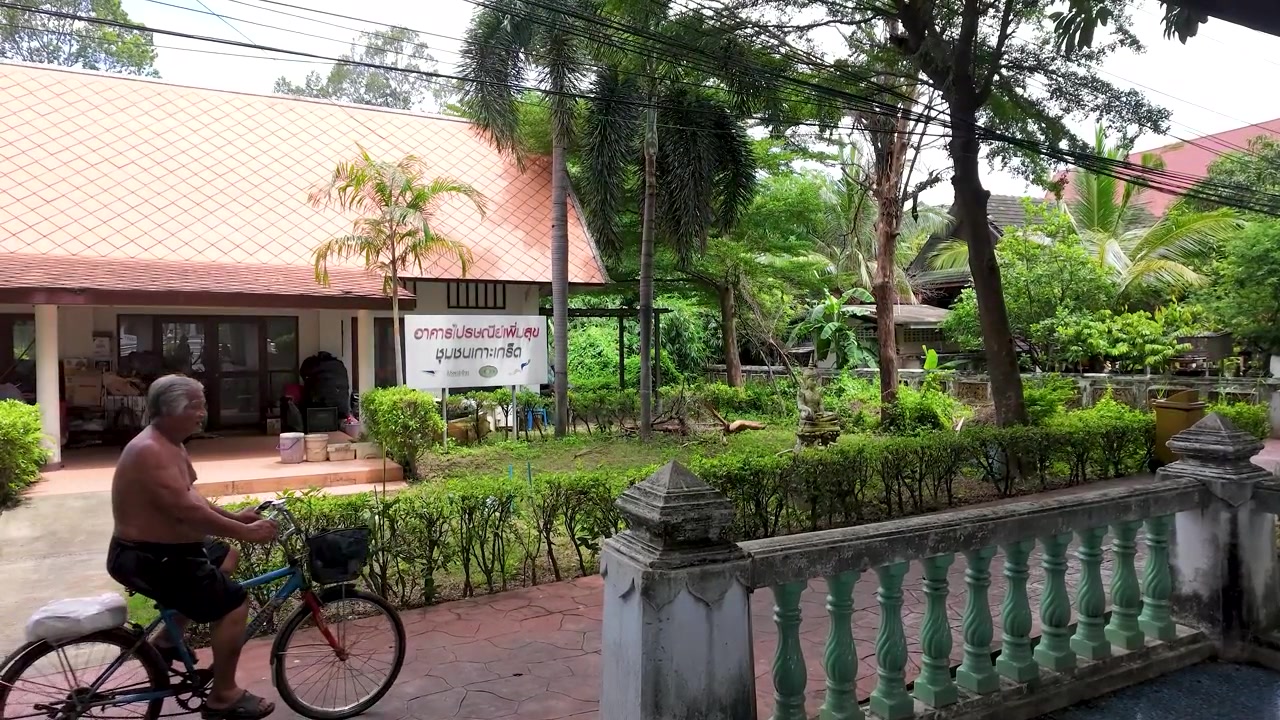 Pria bertelanjang dada bersepeda melewati taman tropis di samping rumah beratap oranye di Thailand.
