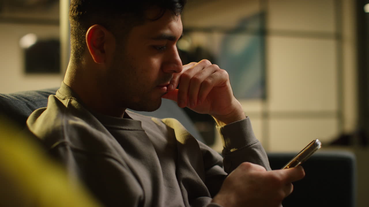 걱정스러운 젊은이가 집에서 저녁을 보내고 소파에 앉아 휴대전화를 들고 인터넷이나 소셜 미디어 스트리밍 콘텐츠를 스크롤합니다.