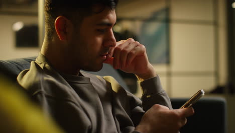 worried young man spending evening at home sitting on sofa with mobile phone scrolling through internet or social media streaming content 1