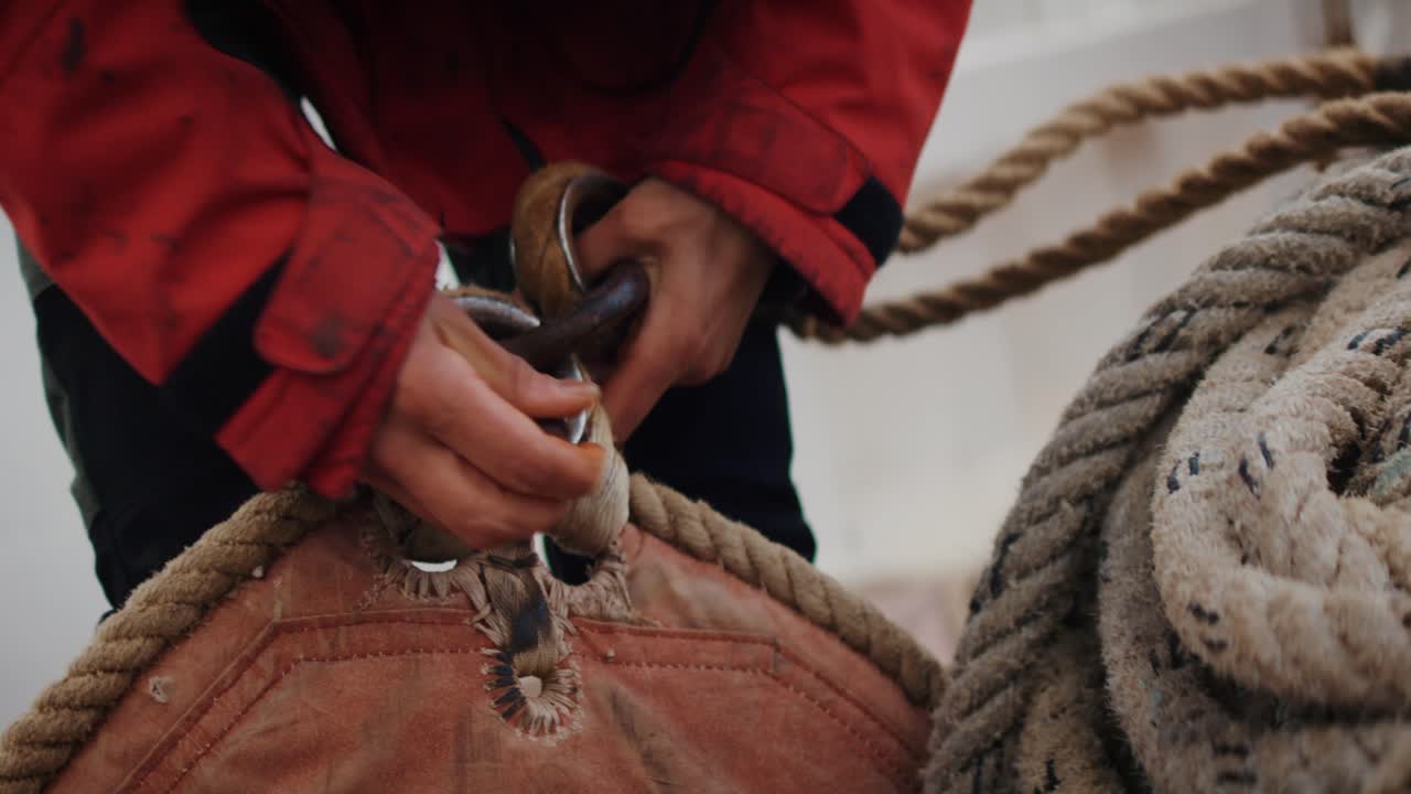 Primo piano di un marinaio che regola l'attrezzatura velica su una barca a vela, mani che stringono una cima e un grillo metallico, dettaglio nautico e concetto di arte marinaresca.