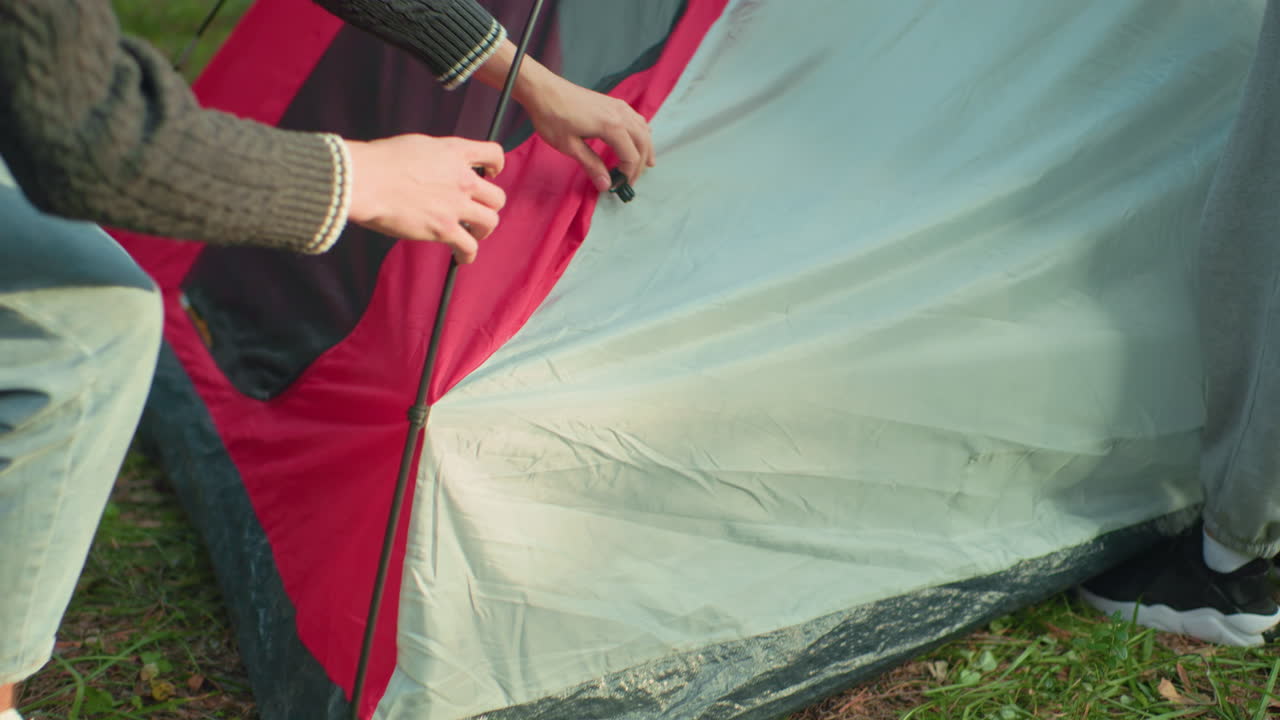 camper crouches fixing tent peg onto flexible pole as sister holds top fabric steady helping secure outdoor shelter together in forest with red and gray tent surrounded by sunlight and green foliage