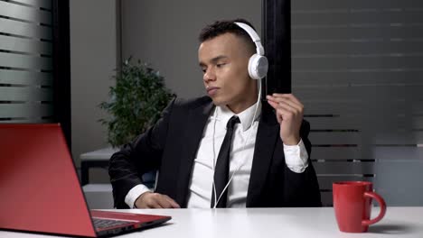 young successful african businessman in suit listening to music on the computer in headphones, dancing in the office. 60 fps