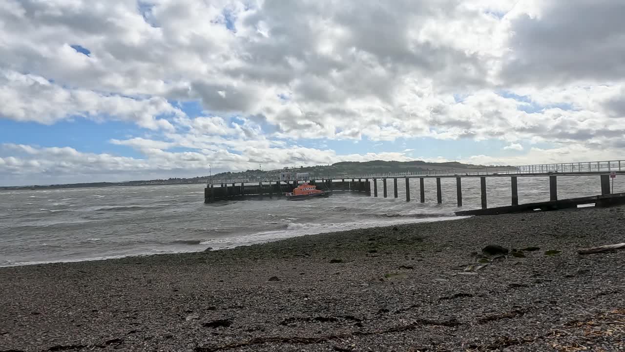 A small orange lifeboat moves toward a wooden pier along a pebbled beach under dramatic, cloudy skies at Broughty Ferry, Scotland. Static wide shot