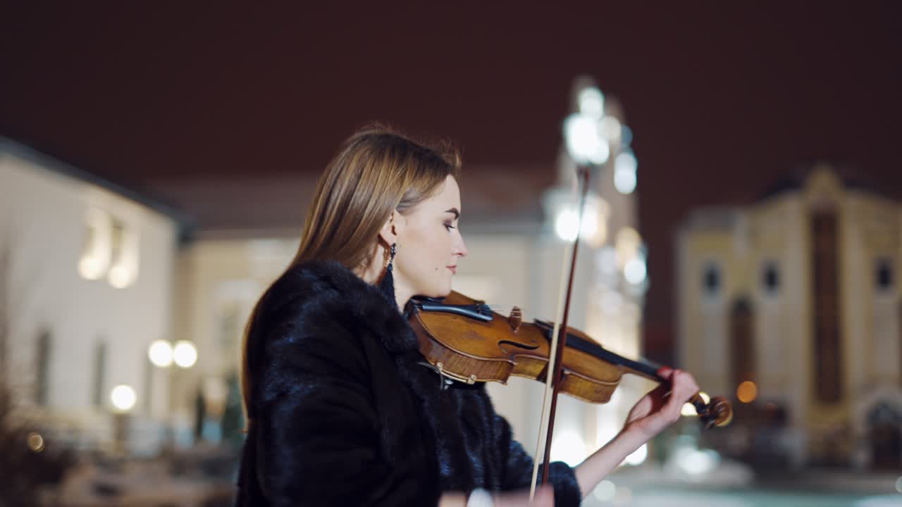 A romantic girl in warm clothes is playing a violin in the street in the square on the background of urban architecture in the evening. Blurred background.