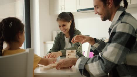 young family playing a game together to develop finger motor skills in a modern kitchen in an apartment. a happy brunette girl in a dark green sweater together with her husband, a brunette guy in a checkered shirt, play with their daughter, a brunette girl in a yellow t-shirt, and assemble a tower o