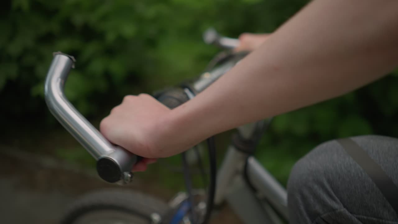 Close-up of cyclist's hand holding the handlebar, riding along a paved path surrounded by lush greenery, light reflects on the cyclist's arm while moving forward with a bokeh background effect