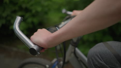 close-up of cyclist's hand holding the handlebar, riding along a paved path surrounded by lush greenery, light reflects on the cyclist's arm while moving forward with a bokeh background effect