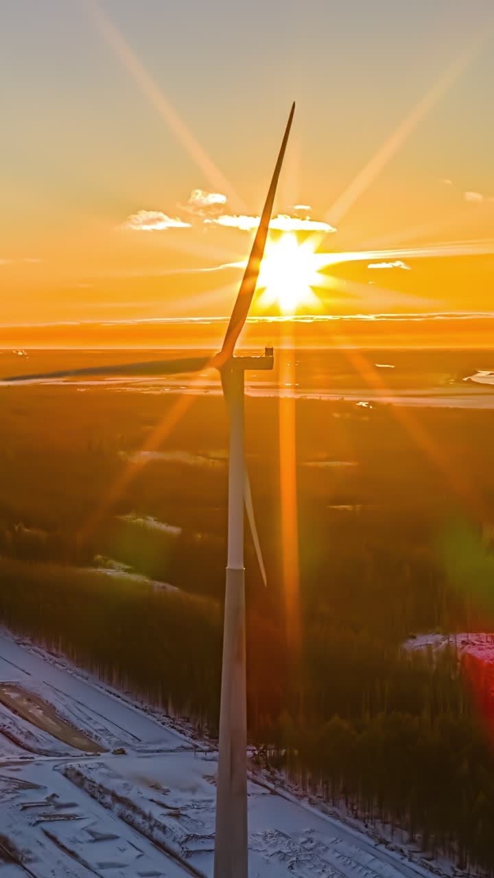 Splendido timelapse aereo verticale di pale di turbine eoliche che ruotano nella luce dorata del tramonto sopra foreste e campi innevati nella Lettonia rurale, con un caldo bagliore arancione e ombre blu