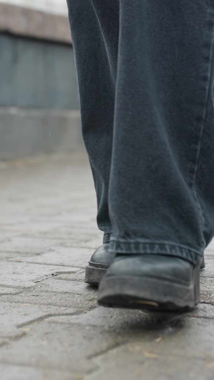 Primo piano dei pantaloni neri e degli stivali neri di una persona che cammina lentamente verso la telecamera su un sentiero urbano pavimentato bagnato, volto non visibile, catturando il dettaglio dei passi, la texture e il movimento silenzioso all'aperto in un freddo autunno.