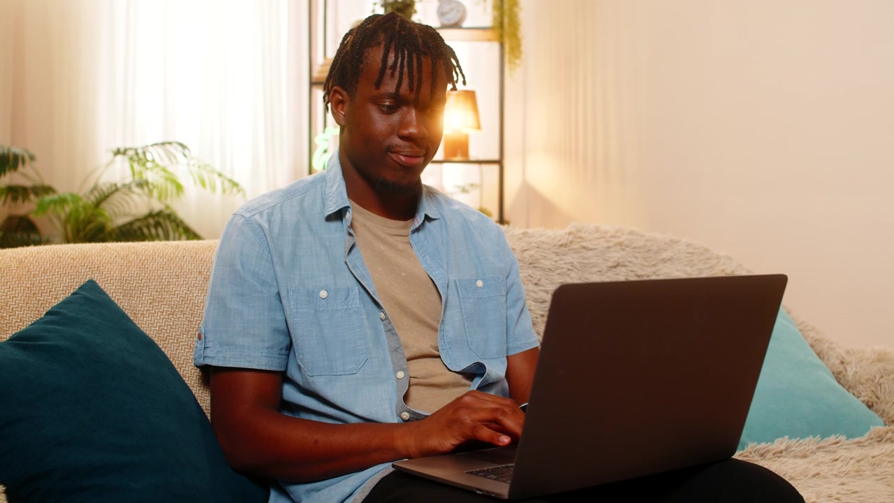 African american young man on home sofa works on laptop reviewing text and correcting small errors