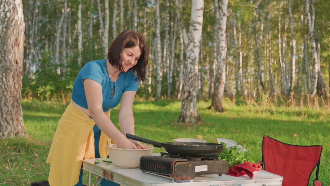 Woman prepping meal on camping stove in sunlit birch forest, folding table with portable gas stove, skillet and fresh vegetables, red camping chair nearby, relaxed picnic atmosphere, casual outdoor
