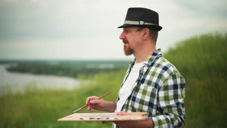 A painter wearing a hat and a checkered shirt stands focused in a serene grassland close to a lake, holding a palette and a paintbrush