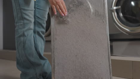 cleaner lifts dusty dryer filter from compartment, sets panel aside, brushes lint buildup, removes debris for safe operation, showing maintenance routine inside modern laundromat, focus on hygiene