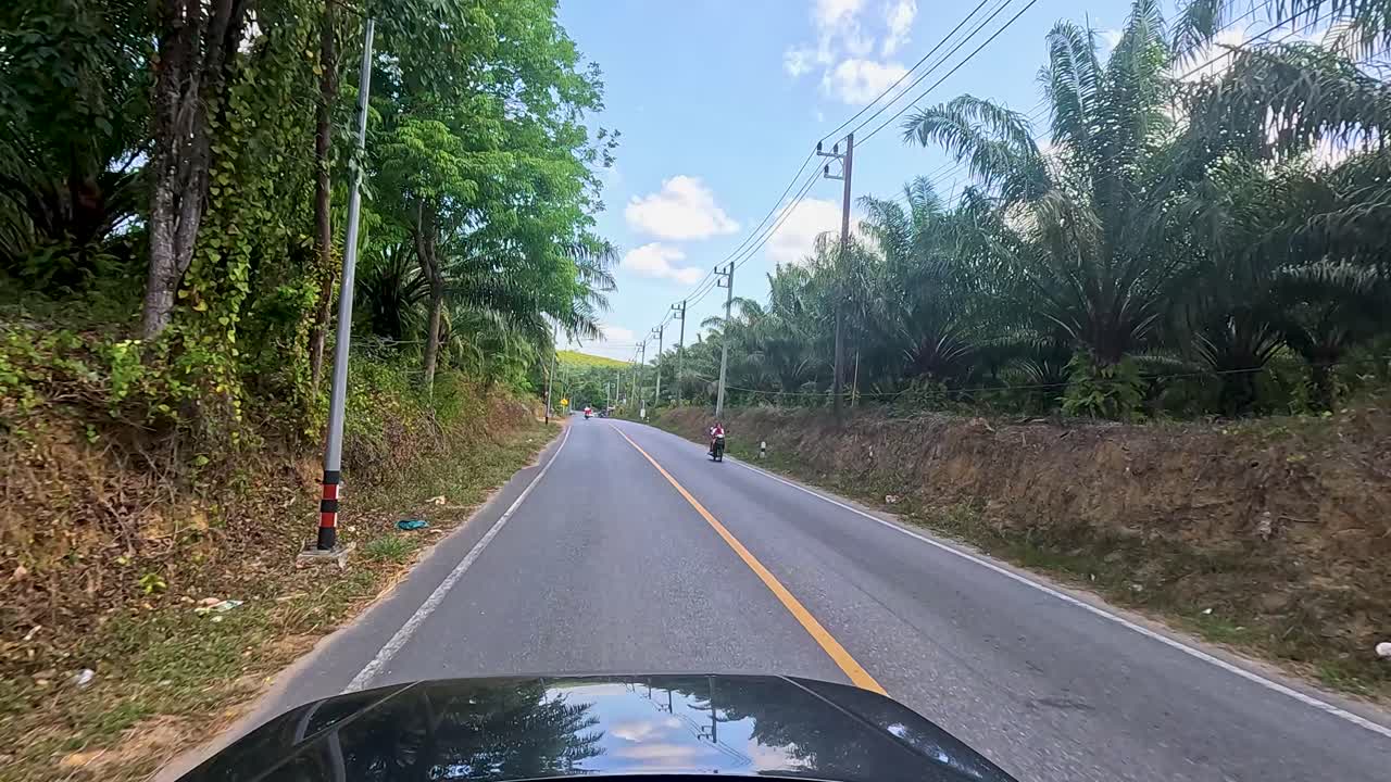 A car drives through a tropical road in Phuket, Thailand, surrounded by lush greenery under a clear blue sky
