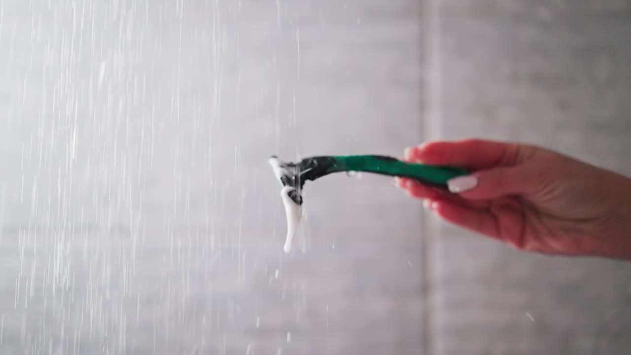 Woman washes razor from foam under shower drops in shower closeup. Lady cleans shaver with water sprays in bathroom. Personal hair removal supplies