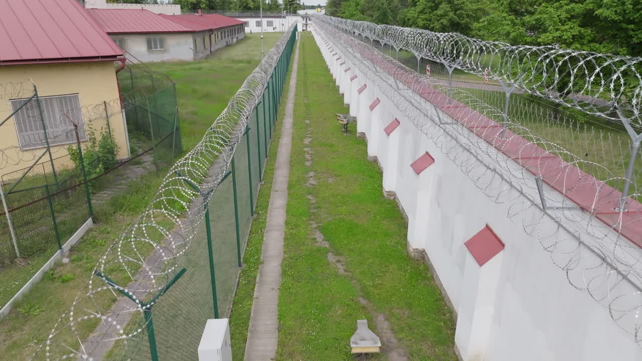 Ripresa a media altitudine con drone che mostra i dettagli di una recinzione di filo spinato, un muro di cemento e sistemi di sicurezza