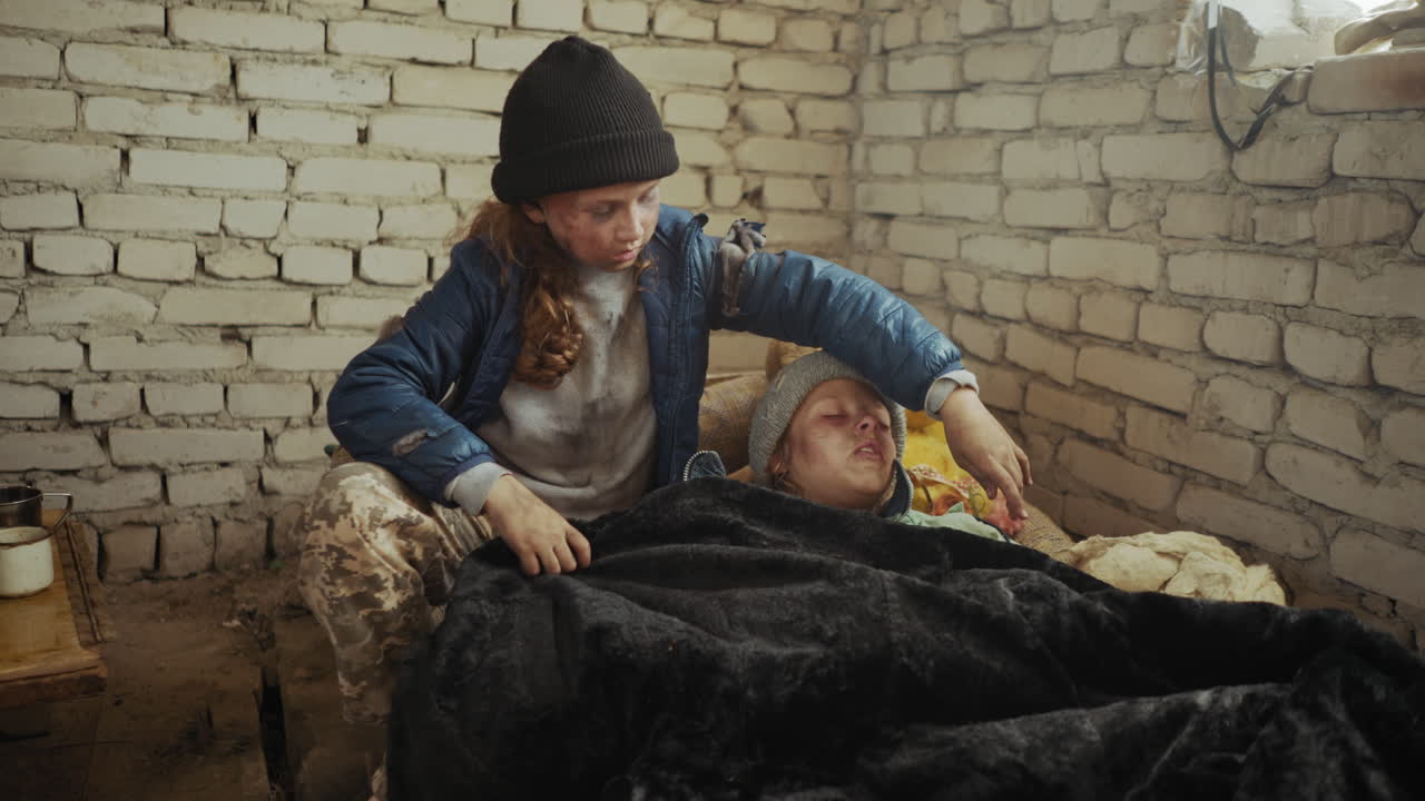 Dirty young girl in winter coat gently covers her sick sibling with blanket inside cold brick shelter, showing compassion, survival instinct, and sibling care in harsh conditions