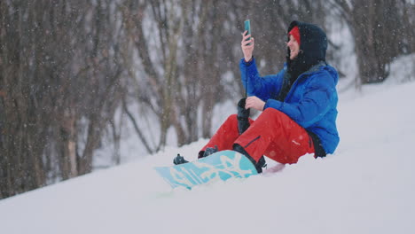 snowboarder masculino sentado en la nieve tomando fotos por teléfono hermosos paisajes del resort para redes sociales