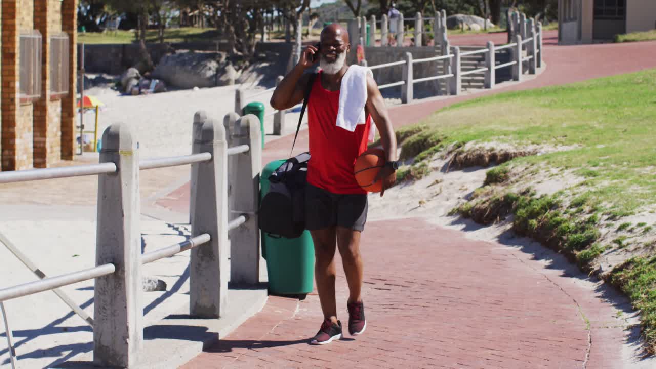 Äldre afroamerikansk man med en basketboll som pratar i smartphone medan han går nära stranden