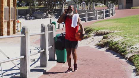 Älterer-Afroamerikanischer-Mann-Mit-Basketball,-Der-Auf-Dem-Smartphone-Spricht,-Während-Er-In-Der-Nähe-Des-Strandes-Spaziert