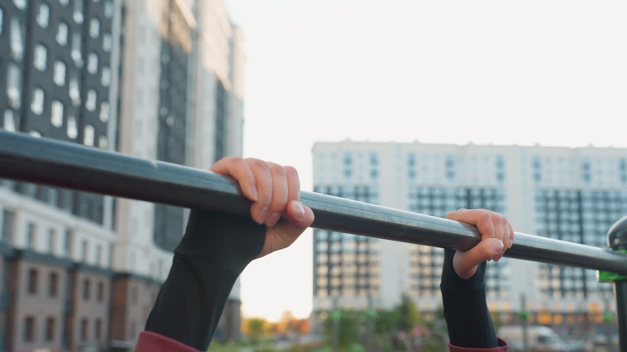 Side hand view of light skinned female exhaling forcefully while gripping horizontal pull up bar in urban park gym setting with city building backdrop showcasing balance and fitness determination