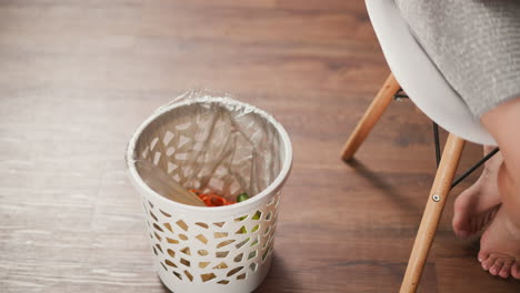 partial view of young adult seated on white chair inspecting cucumber and disposing it into waste bin lined with plastic bag on wooden floor indoor kitchen