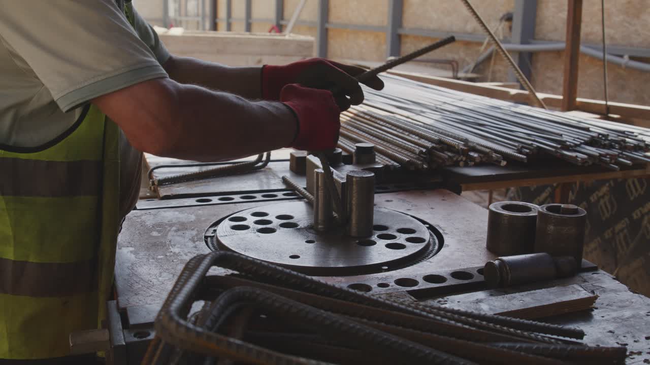 A worker is seen using a machine to bend steel rebars on a construction site. The worker is wearing protective gloves and a high-visibility vest